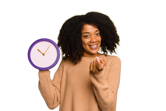 Young African American holding a clock isolated pointing with finger at you as if inviting come closer.