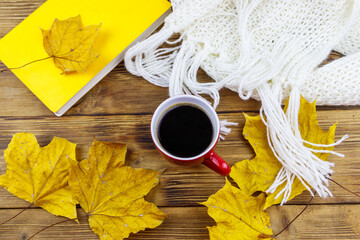 Knitted white scarf, book, cup of coffee and autumn maple leaves on wooden table. Top view. Autumn cozy concept