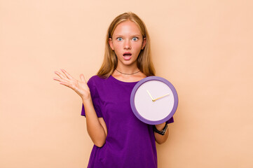 Little caucasian girl holding a clock isolated on beige background surprised and shocked.