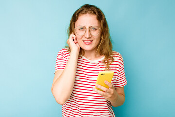 Young caucasian woman holding mobile phone isolated on blue background covering ears with hands.