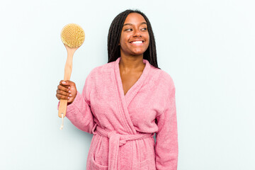 Young African American woman holding a bathtub brush isolated on blue background looks aside smiling, cheerful and pleasant.