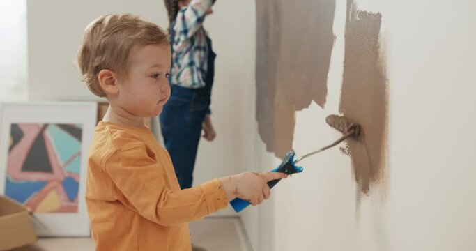 Little boy in orange sweater holds roller in hands and paints the wall brown. The boy has blond hair, looking at sister, who is drawing from the side. Girl with pigtails looks at brother and smiles.