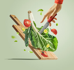 Women hand with knife , flying salad ingredients and cutting board at light green background....