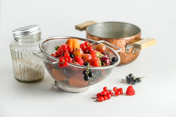 Jam ingredients: various fruits and berries in colander with cooking pot and sugar in glass jar standing on white background