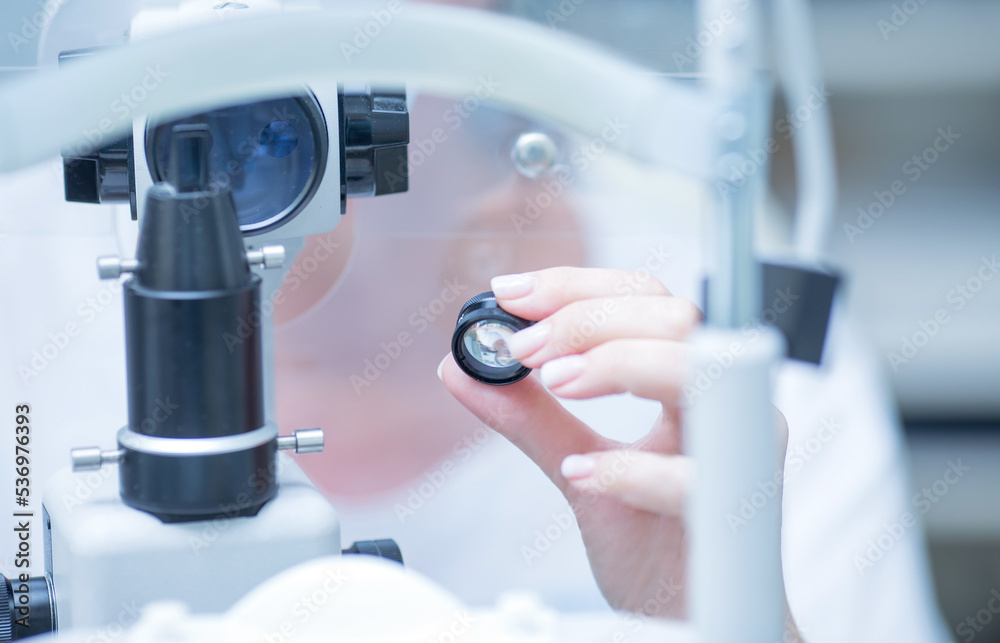 Wall mural a young ophthalmologist woman looks into the viewfinder of a slit lamp for biomicroscopy of the eye. in the hand is a lens for examining thefundus .