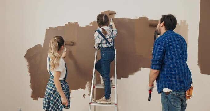 Young family paints walls in the living room in brown. They stand with backs to the camera, holding rollers and wearing checkered shirts. Girl with pigtails in denim overalls is standing on ladder.