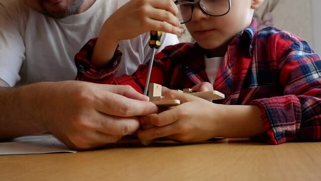 Happy father's day and childhood concept. Close-up of a father and a boy son in glasses work with hand tools, using a screwdriver, assembling a wooden house constructor at the table.