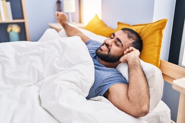 Young hispanic man waking up stretching arms at bedroom