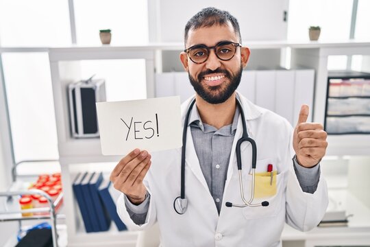 Middle east man with beard wearing doctor uniform and stethoscope holding yes banner smiling happy and positive, thumb up doing excellent and approval sign