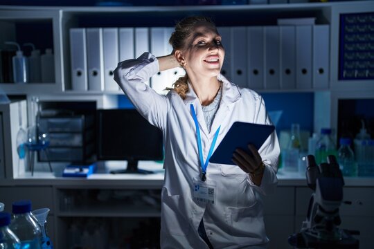 Beautiful blonde woman working at scientist laboratory late at night smiling confident touching hair with hand up gesture, posing attractive and fashionable