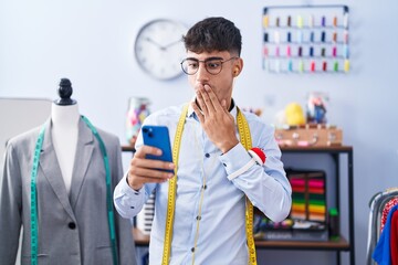 Young hispanic man dressmaker designer using smartphone covering mouth with hand, shocked and afraid for mistake. surprised expression
