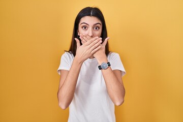Young beautiful woman standing over yellow background shocked covering mouth with hands for...
