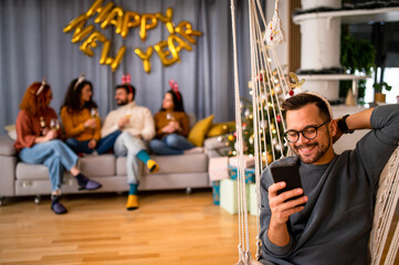 Friends having fun at a Christmas party in their decorated apartment, wearing festive sweaters, chatting and scrolling through their phone 