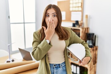 Young brunette woman holding architect hardhat at the office covering mouth with hand, shocked and...