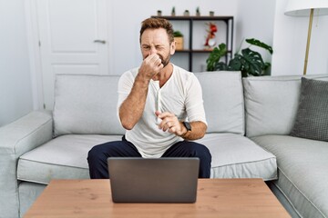 Middle age man using laptop at home smelling something stinky and disgusting, intolerable smell, holding breath with fingers on nose. bad smell - obrazy, fototapety, plakaty