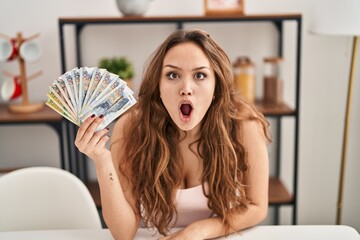 Young hispanic woman holding peruvian sol banknotes scared and amazed with open mouth for surprise, disbelief face - obrazy, fototapety, plakaty