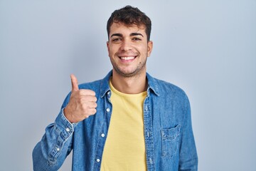 Young hispanic man standing over blue background doing happy thumbs up gesture with hand. approving expression looking at the camera showing success.