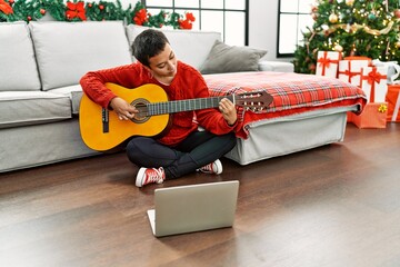 Young hispanic woman having online guitar class sitting by christmas tree at home