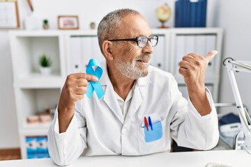 Mature doctor man holding blue ribbon at the clinic pointing thumb up to the side smiling happy with open mouth
