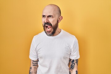 Young hispanic man with tattoos standing over yellow background angry and mad screaming frustrated and furious, shouting with anger. rage and aggressive concept.