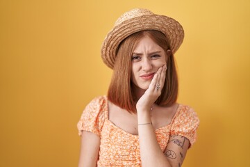 Young redhead woman standing over yellow background wearing summer hat touching mouth with hand with painful expression because of toothache or dental illness on teeth. dentist