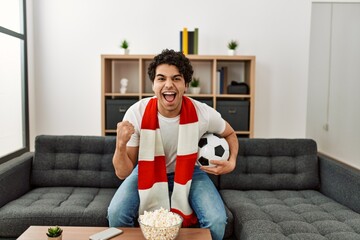 Young hispanic man watching soccer match and supporting team at home.