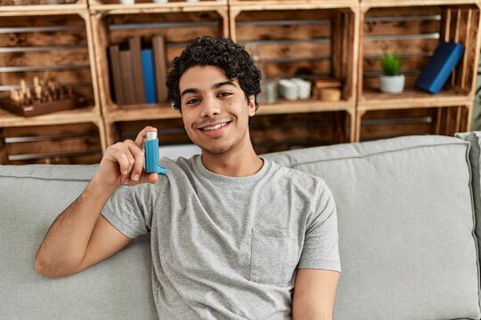 Young hispanic man using inhaler sitting on the sofa at home.
