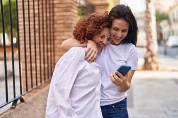 Two women mother and daughter hugging each other using smartphone at street