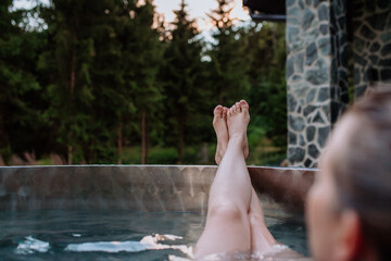 Unrecognizable young woman with feet up relaxing in hot tub outdoor in nature.
