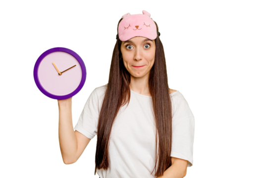 Young caucasian woman wearing pajama and holding a clock shrugs shoulders and open eyes confused.