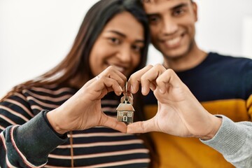 Young latin couple doing heart symbol with hands holding key of new home.