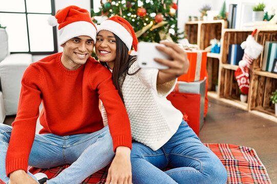 Young latin couple smiling happy making selfie by the smartphone sitting on the floor by christmas tree at home.