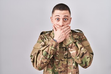 Young man wearing camouflage army uniform shocked covering mouth with hands for mistake. secret...