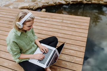 Woman, freelancer working on laptop, sitting on a pier by the backyard lake, a concept of remote office, work during vacation.
