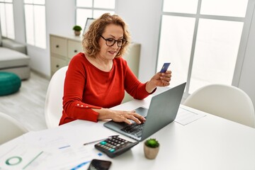 Middle age caucasian woman using laptop and credit card sitting on the table at home.