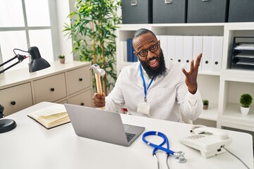 African american man working at medical clinic holding hammer celebrating victory with happy smile and winner expression with raised hands