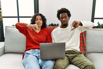 Young interracial couple sitting on the sofa on christmas using laptop with angry face, negative...