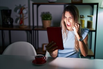 Young hispanic woman using touchpad sitting on the table at night angry and mad raising fist frustrated and furious while shouting with anger. rage and aggressive concept.