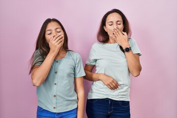 Young mother and daughter standing over pink background bored yawning tired covering mouth with...