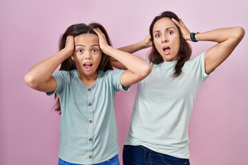 Young mother and daughter standing over pink background crazy and scared with hands on head, afraid...