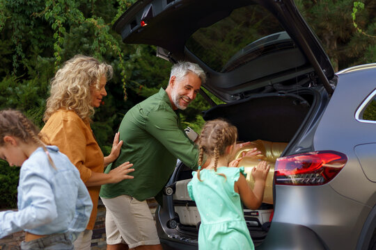 Happy family preparing for holiday, putting suitcases in car trunk, while their electric car charging.