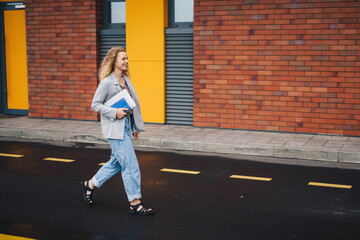 Side view of a woman student wearing jeans and jacket holding laptop computer confidently walking on street in afternoon. Cheerful student or a businesswoman.