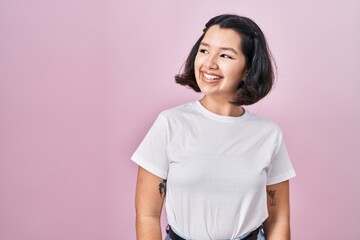 Young hispanic woman wearing casual white t shirt over pink background looking away to side with smile on face, natural expression. laughing confident.