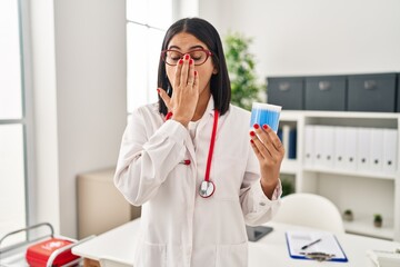 Young hispanic doctor woman holding cotton buds covering mouth with hand, shocked and afraid for...