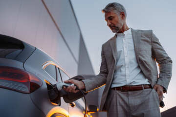 Businessman, holding power supply and charging his electric car during sunset. Concept of ecology...