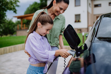 Happy mother showing her little daughter how to charge their electric car.