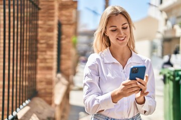 Young blonde woman smiling confident using smartphone at street