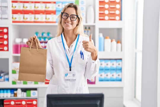Young blonde woman working at pharmacy drugstore holding paper bag smiling happy and positive, thumb up doing excellent and approval sign