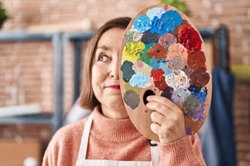 Middle age hispanic woman holding painter palette close to face smiling looking to the side and...