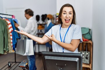 Middle age hispanic woman working as manager at retail boutique amazed and smiling to the camera while presenting with hand and pointing with finger.